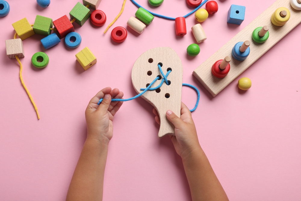Motor skills development. Little child playing with wooden lacing toy at pink table, top view